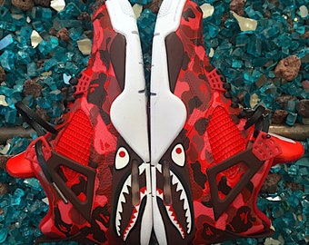May include: A pair of red and black sneakers with a camouflage pattern and shark teeth design. The sneakers are on a background of blue and brown rocks.
