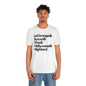 以下が含まれることがあります： 白のTシャツに、LaCienega、Sunset、Vine、Hollywood、Highlandのストリート名を黒字で記載。Tシャツは半袖で柔らかい素材。人物はブルージーンズを着用しています。
