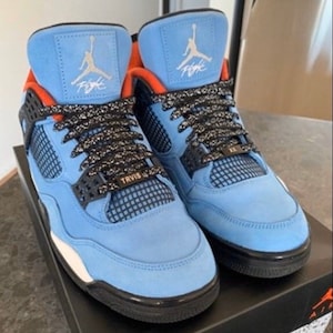 Puede incluir: Un par de zapatillas de gamuza azul claro con detalles en negro y naranja. Las zapatillas tienen un panel de malla negra en el lateral y una entresuela blanca. El texto "TRAVIS SCOTT" est&aacute; impreso en el lateral de las zapatillas.