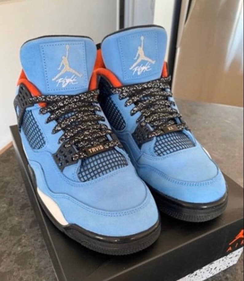 Puede incluir: Un par de zapatillas de gamuza azul claro con detalles en negro y naranja. Las zapatillas tienen un panel de malla negra en el lateral y una entresuela blanca. El texto "TRAVIS SCOTT" est&aacute; impreso en el lateral de las zapatillas.