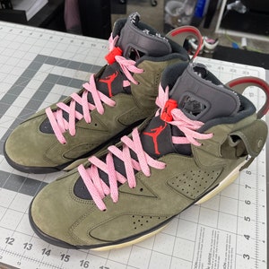 May include: A pair of olive green and black high-top sneakers with pink laces. The sneakers feature a red Jumpman logo and a gray tongue. The soles are off-white and the shoes are on a gray and white grid surface.