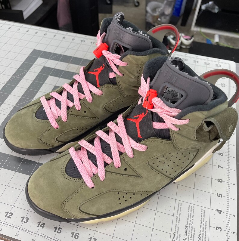 May include: A pair of olive green and black high-top sneakers with pink laces. The sneakers feature a red Jumpman logo and a gray tongue. The soles are off-white and the shoes are on a gray and white grid surface.