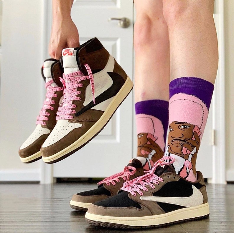 Puede incluir: Dos pares de zapatillas deportivas, una de ca&ntilde;a alta y otra baja, en marr&oacute;n, crema y negro, con cordones rosas. Las zapatillas de ca&ntilde;a alta son sostenidas por una persona. La persona lleva calcetines morados y rosas con un dibujo animado.