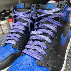 May include: Close-up of a pair of high-top sneakers. The shoes feature a black and royal blue color scheme with purple laces. The Nike Air logo is visible on the tongue. The shoes are on a cutting mat.