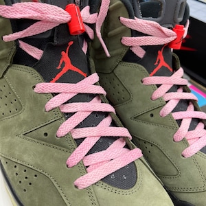 May include: Close-up of olive green and black high-top sneakers with pink laces. The sneakers feature the Jumpman logo in red on the tongue and side. The laces are threaded through the eyelets and secured with red lace locks.