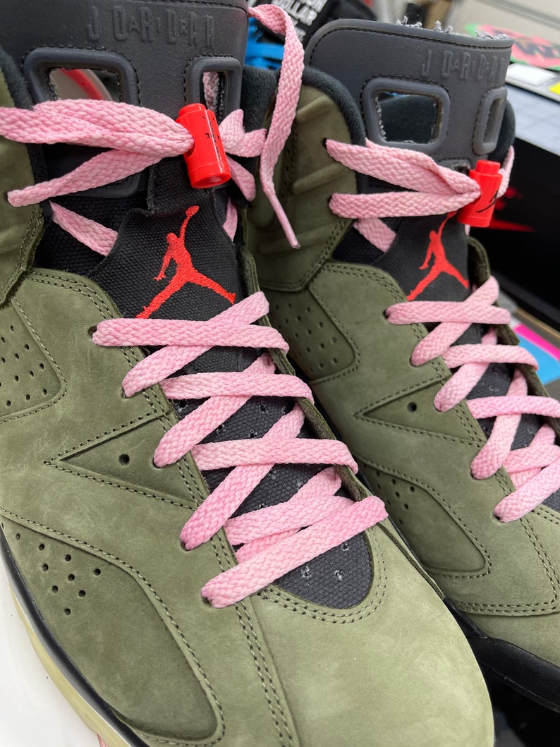 May include: Close-up of olive green and black high-top sneakers with pink laces. The sneakers feature the Jumpman logo in red on the tongue and side. The laces are threaded through the eyelets and secured with red lace locks.