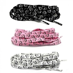 Puede incluir: Tres pares de cordones en negro, rosa y blanco. Cada par presenta un patr&oacute;n repetido de una cara estilizada en negro. Los cordones est&aacute;n cuidadosamente enrollados, mostrando sus dise&ntilde;os y variaciones de color.