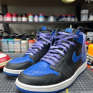 May include: Black and blue high-top sneakers with purple laces. The sneakers have a blue toe box and a blue Nike swoosh. The midsole is white, and the outsole is blue. The word "AIR" is visible on the tongue.