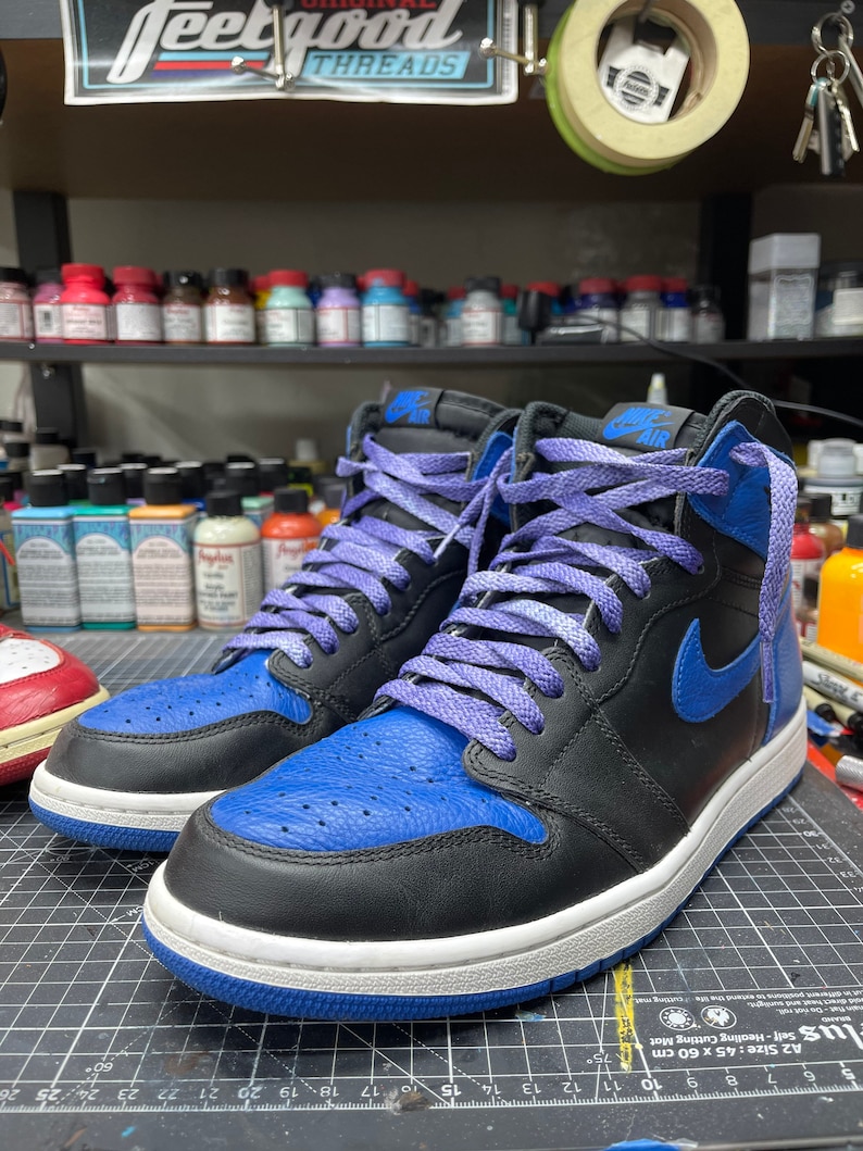 May include: Black and blue high-top sneakers with purple laces. The sneakers have a blue toe box and a blue Nike swoosh. The midsole is white, and the outsole is blue. The word "AIR" is visible on the tongue.