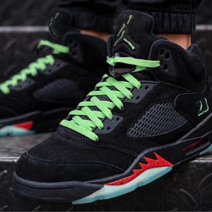 May include: Black high-top sneakers featuring bright green laces, a translucent lace lock, and a black suede upper. The midsole includes a red shark-tooth design with black speckles, and the outsole is a translucent light blue material.