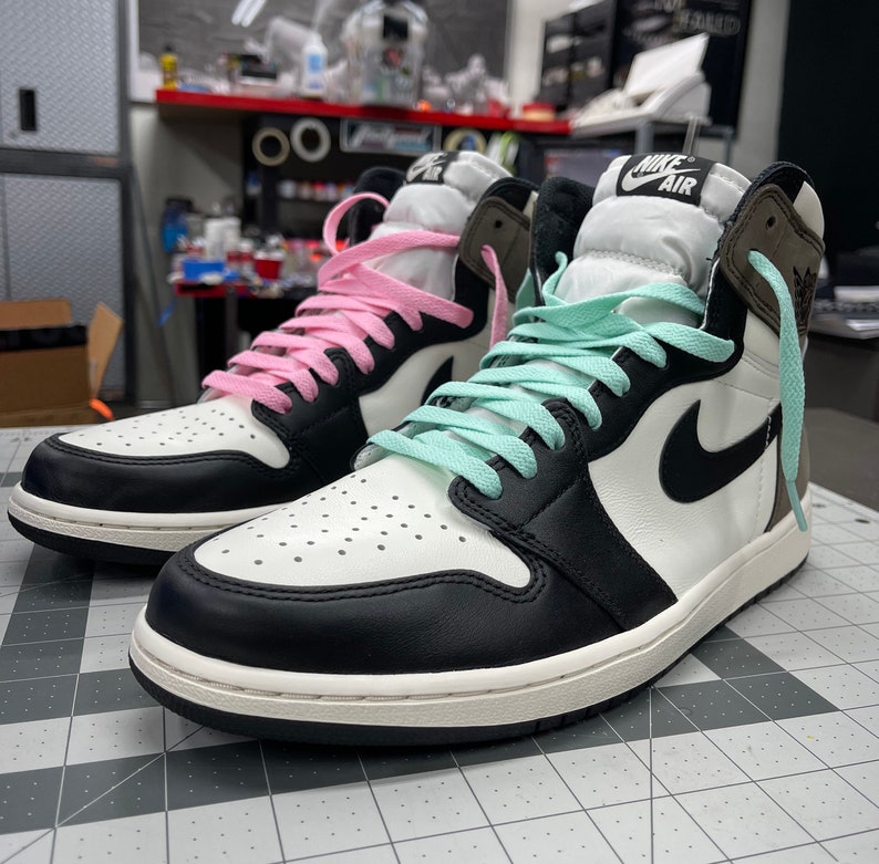 May include: A pair of black and white sneakers with a light brown suede overlay. One shoe has pink laces and the other has light blue laces. The shoes have the Nike Air logo on the side.