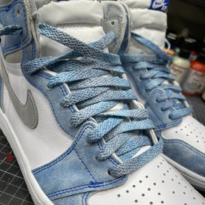 May include: Close-up of a pair of high-top sneakers. The shoes feature a white leather base with light blue suede overlays and laces. A gray Nike swoosh is visible on the side. The tongue has a blue tag with "NIKE AIR" in white.