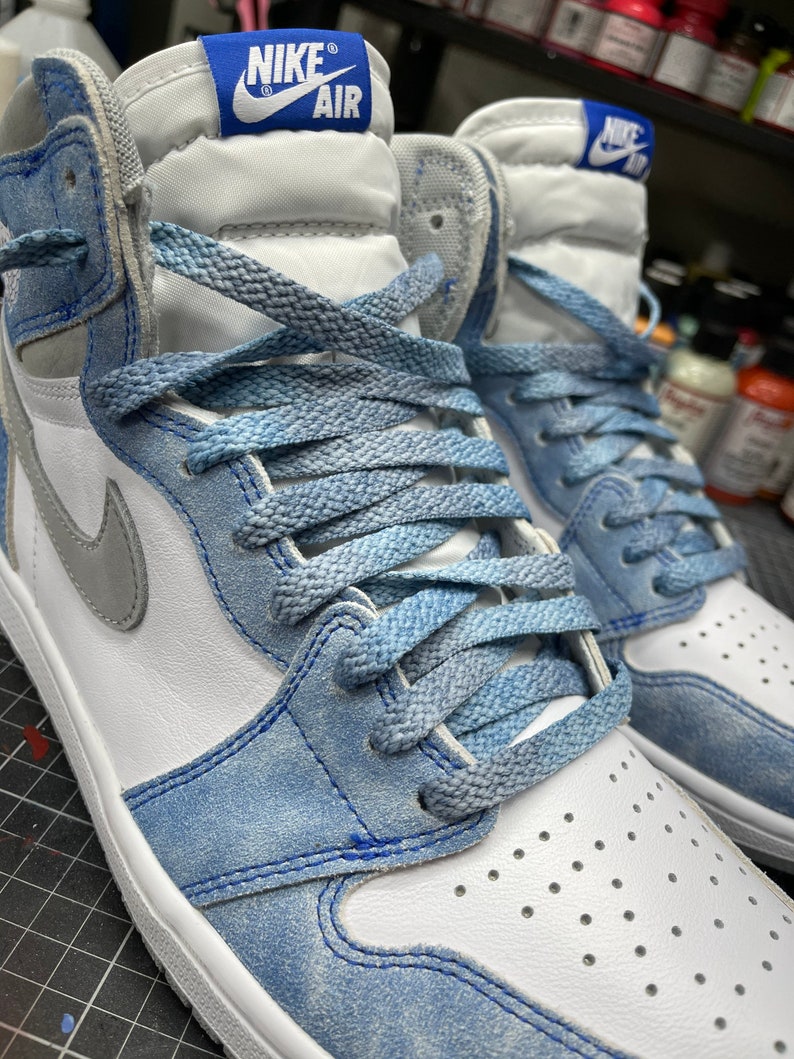 May include: Close-up of a pair of high-top sneakers. The shoes feature a white leather base with light blue suede overlays and laces. A gray Nike swoosh is visible on the side. The tongue has a blue tag with "NIKE AIR" in white.