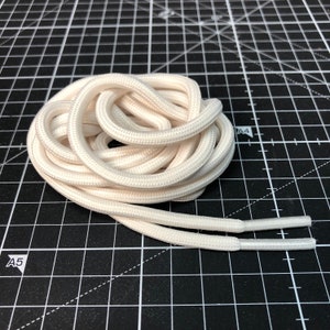 Puede incluir: Un par de cordones blancos, enrollados y dispuestos sobre una superficie de rejilla negra.