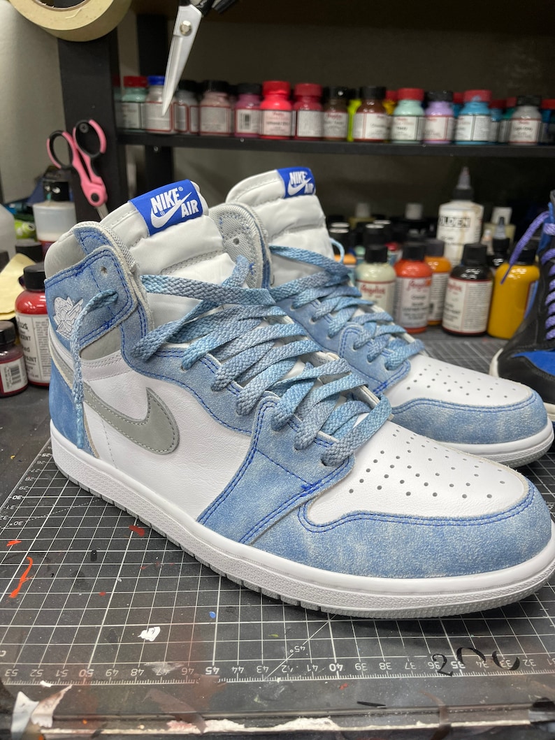 May include: Pair of high-top sneakers with white, light blue, and gray accents. The sneakers feature the Nike Air logo on the tongue and a gray Nike swoosh. The laces are light blue, and the soles are white.