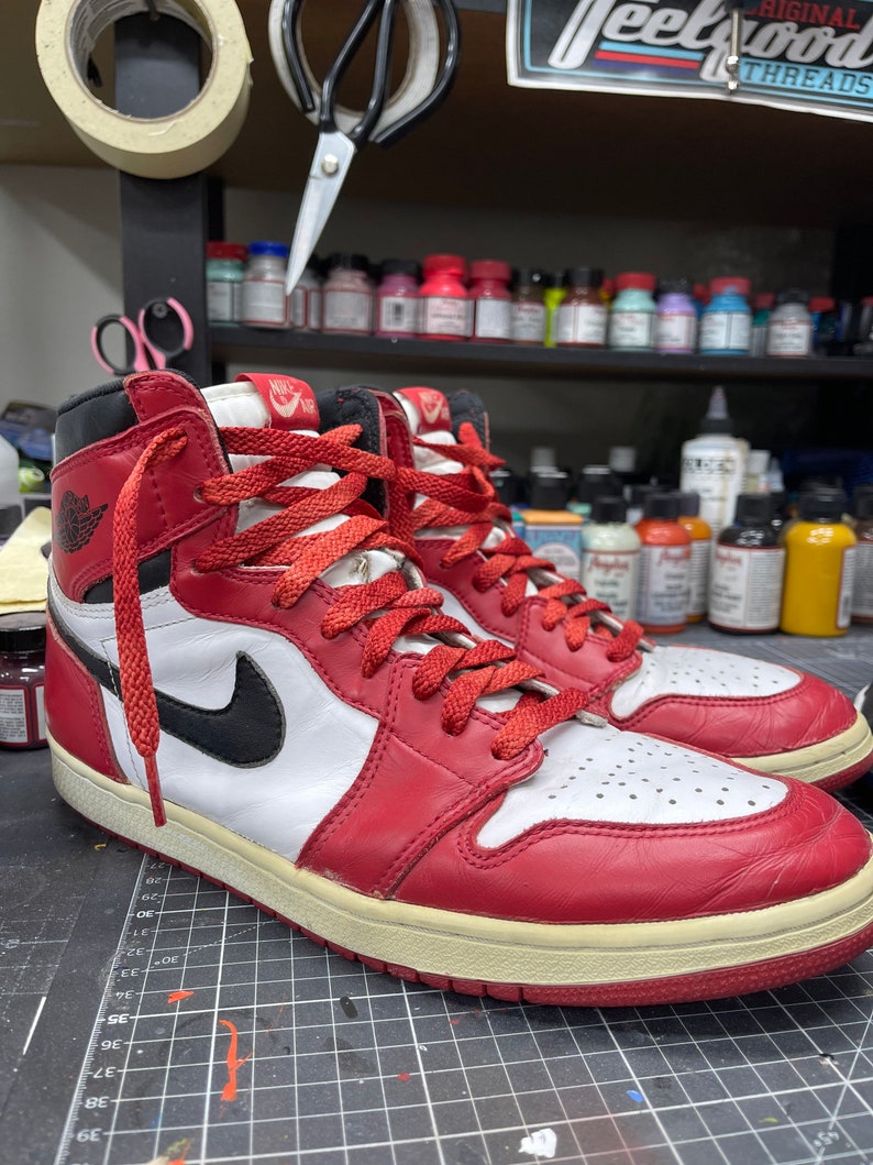 May include: A pair of high-top sneakers in red, white, and black. The sneakers feature a red leather upper with white panels, a black swoosh, and red laces. The soles are off-white. The sneakers are on a black surface.