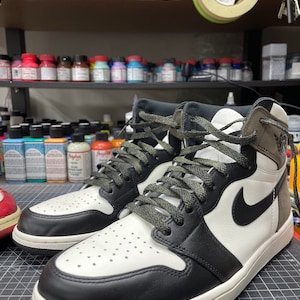 May include: A pair of high-top sneakers with a black and white color scheme. The sneakers feature a black Nike swoosh, black laces, and a white midsole. The uppers are a combination of black, white, and brown leather. The sneakers are displayed on a cutting mat.