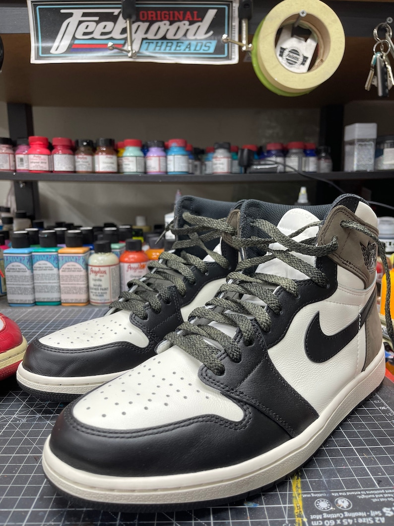 May include: A pair of high-top sneakers with a black and white color scheme. The sneakers feature a black Nike swoosh, black laces, and a white midsole. The uppers are a combination of black, white, and brown leather. The sneakers are displayed on a cutting mat.