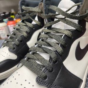 May include: Close-up of a pair of high-top sneakers. The shoes feature a white leather upper with black leather overlays and a black Nike swoosh. The laces are olive green, and the tongue and collar are dark gray.