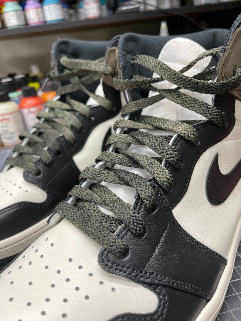 May include: Close-up of a pair of high-top sneakers. The shoes feature a white leather upper with black leather overlays and a black Nike swoosh. The laces are olive green, and the tongue and collar are dark gray.