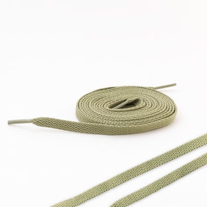 May include: A pair of sage green shoelaces with a textured surface. The shoelaces are laid out flat on a white background.