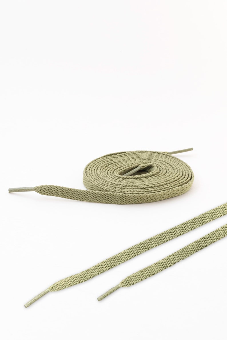 May include: A pair of sage green shoelaces with a textured surface. The shoelaces are laid out flat on a white background.