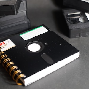 May include: A black 3.5 inch floppy disk notebook with a gold spiral binding. The disk has a white label with the text "3M DS/HD" printed on it. The notebook is sitting on a gray surface.