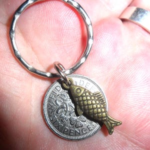 May include: A silver sixpence coin keychain with a small gold-coloured fish charm attached. The coin has the inscription "SIXPENCE" and a floral design.