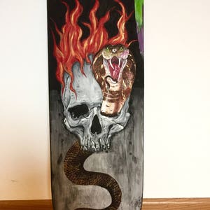 May include: A hand-painted skateboard deck with a skull and a cobra snake. The snake is wrapped around the skull and has its mouth open. The skull is on fire. The background is black and gray.