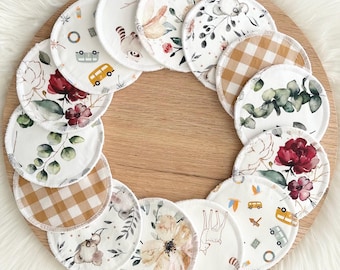 May include: A set of 14 round, white cotton makeup remover pads with various patterns. The patterns include floral, geometric, and animal prints. The pads are arranged in a circle on a wooden surface.