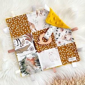May include: A multi-textured baby sensory blanket in yellow, white and brown. The blanket features various animal and nature-themed patches, including a kangaroo, a crocodile, a bear and a floral design. The blanket also includes a wooden teether and a tag with the text "Wild & Free".