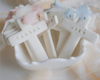 May include: A collection of white ceramic crosses with stamped names including TAMARA and JOSE, each adorned with a soft pastel pink or light blue fabric ribbon tie, arranged together in a small white scalloped bowl for display.