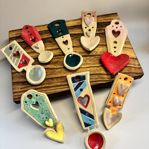 Puede incluir: Una colección de cucharas de cerámica hechas a mano con recortes en forma de corazón y elementos decorativos. Las cucharas presentan varios colores, incluyendo rojo, azul, verde y amarillo, y están dispuestas sobre una tabla de madera. Cada cuchara tiene un diseño y forma únicos.