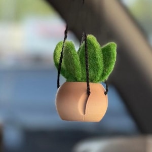 May include: A hanging car ornament featuring a small, green, felted succulent plant potted in a rounded, peach-colored container. The plant is suspended by thin black strings attached to the top of the vehicle's rearview mirror assembly.