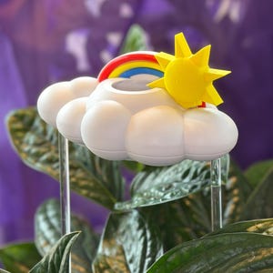 K&ouml;nnte beinhalten: Ein wei&szlig;er, wolkenf&ouml;rmiger Pflanzenstecker mit einer leuchtend gelben Sonne und einem bunten Regenbogen auf der Oberseite. Das Dekorationsobjekt wird von zwei transparenten Acrylst&auml;ben gehalten, die von der Unterseite der Wolke nach unten ragen.
