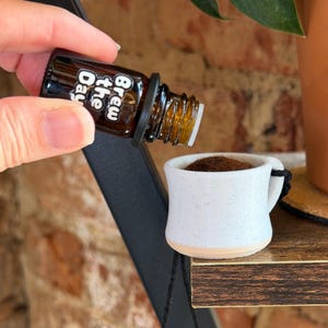 May include: A small brown bottle labeled "Brew the Day" is pouring into a miniature white mug filled with brown granules. The mug has a black handle and a beige base. The scene is set on a wooden shelf.