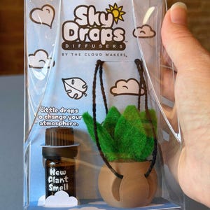 May include: A Sky Drops Diffuser kit in clear plastic packaging containing a small brown glass bottle labeled New Plant Smell and a miniature green felt plant in a tan pot with black hanging strings. The packaging features clouds and text.