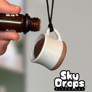 Pode incluir: Um difusor de óleos essenciais em forma de chávena de café de cerâmica branca e castanha, pendurado por um cordão preto. Um frasco de óleo essencial castanho está a verter óleo para o difusor. O logótipo Sky Drops Diffusers está no canto inferior direito.