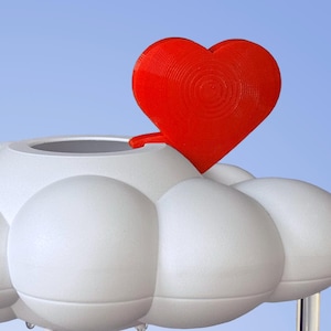 Puede incluir: Un macetero blanco en forma de nube con una tapa roja en forma de corazón. El macetero tiene un agujero en la parte superior para el drenaje.