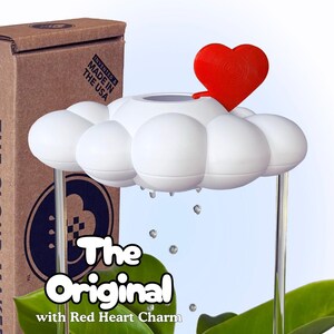 Puede incluir: Un dispositivo de riego para plantas con forma de nube blanca y un dije de corazón rojo en la parte superior. El objeto tiene soportes verticales transparentes sobre hojas verdes, con pequeñas gotas transparentes suspendidas. La caja dice The Original with Red Heart Charm.