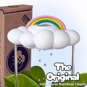 Peut inclure: Un objet décoratif blanc en forme de nuage avec un accent arc-en-ciel pastel. L'objet a des tiges transparentes et est positionné devant des feuilles vertes. Le texte "The Original with Pastel Rainbow Charm" est affiché sur l'image.