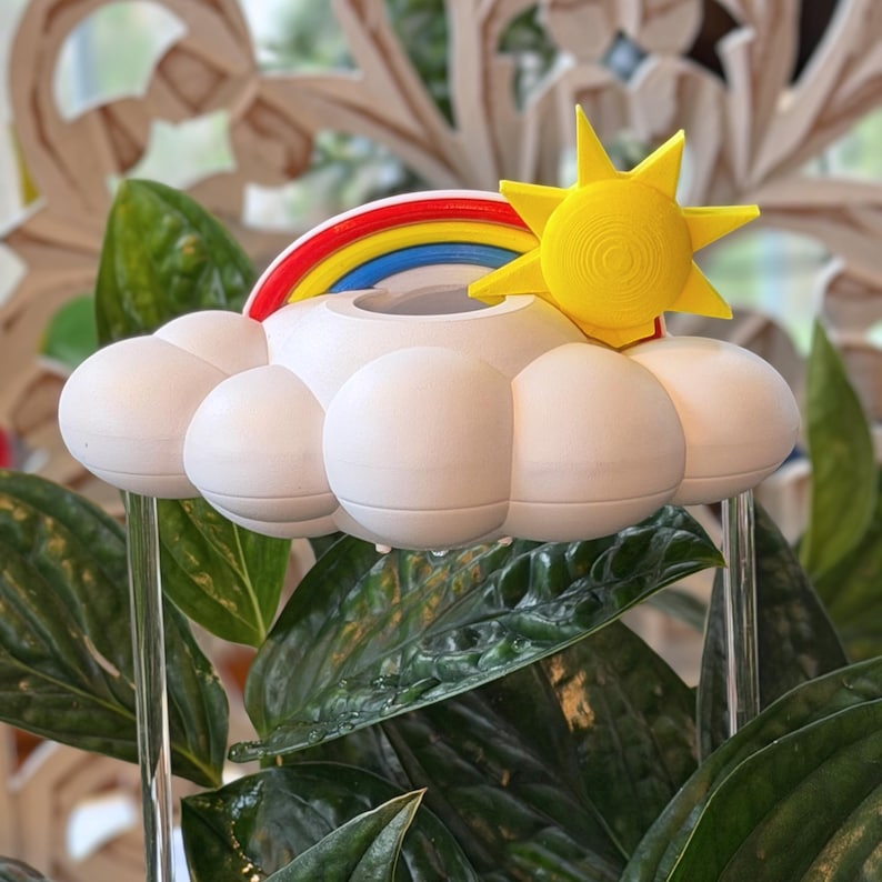 K&ouml;nnte beinhalten: Eine wei&szlig;e, wolkenf&ouml;rmige Pflanzenbew&auml;sserungshilfe mit einer leuchtend gelben Sonne und einem bunten Regenbogenbogen. Die Oberseite der Wolke hat eine kreisf&ouml;rmige &Ouml;ffnung f&uuml;r Wasser und wird von zwei klaren, vertikalen St&auml;ben gest&uuml;tzt.