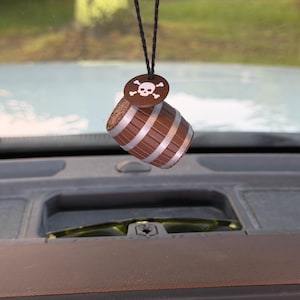 May include: A brown barrel-shaped hanging ornament featuring silver bands and a circular brown tag at the top with a white skull and crossbones graphic. The object is suspended by a thin black cord, displaying a textured wood-like finish.