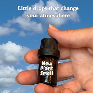 May include: A small amber glass bottle with a black cap and a white label that reads "New Plant Smell". Above the bottle, white text reads "Little drops that change your atmosphere". The bottle is held against a bright blue sky with clouds.