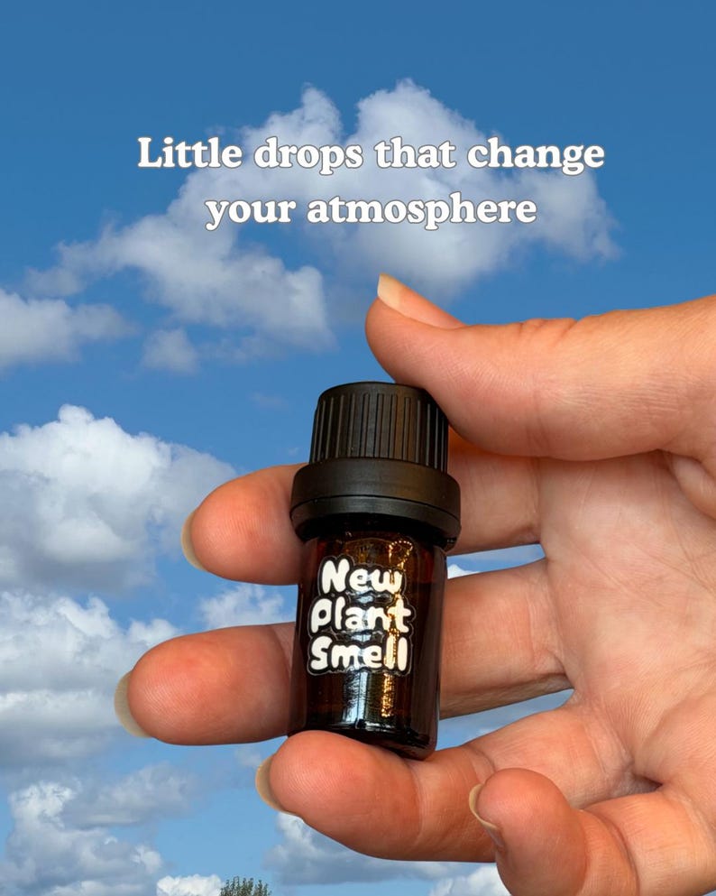 May include: A small amber glass bottle with a black cap and a white label that reads "New Plant Smell". Above the bottle, white text reads "Little drops that change your atmosphere". The bottle is held against a bright blue sky with clouds.