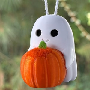 May include: A white ghost ornament with black circular eyes and a small smile, holding a bright orange pumpkin with a green stem. The ghost is suspended by a thin white string attached to the top of its head, creating a hanging decoration.