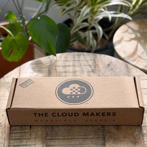 K&ouml;nnte beinhalten: Ein rechteckiger brauner Karton mit einem schwarzen kreisf&ouml;rmigen Logo, das eine Wolke und Regentropfen zeigt. Der Text lautet THE CLOUD MAKERS WOODSTOCK, GEORGIA und INVENTED & MADE IN THE USA. Die Schachtel hat eine flache Oberseite und Beschriftung.