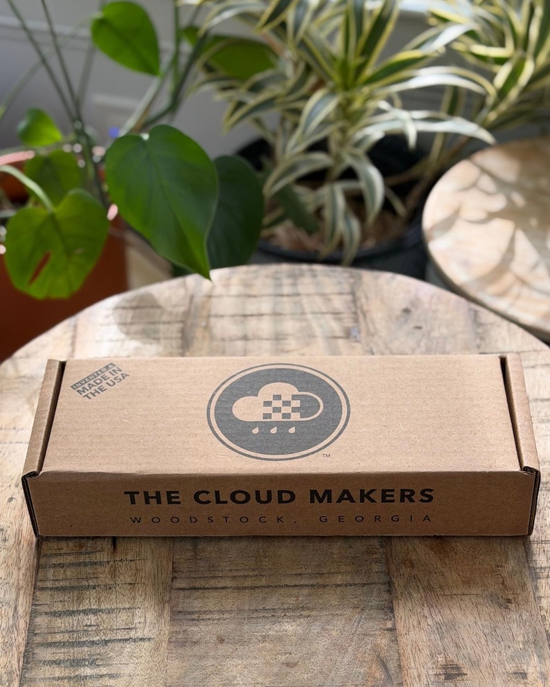 K&ouml;nnte beinhalten: Ein rechteckiger brauner Karton mit einem schwarzen kreisf&ouml;rmigen Logo, das eine Wolke und Regentropfen zeigt. Der Text lautet THE CLOUD MAKERS WOODSTOCK, GEORGIA und INVENTED & MADE IN THE USA. Die Schachtel hat eine flache Oberseite und Beschriftung.