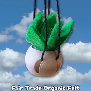 May include: A hanging plant decoration featuring bright green felt leaves arranged in a rounded, tan-colored planter. The planter is suspended by three black cords. The text "Fair Trade Organic Felt" is printed at the bottom of the image.