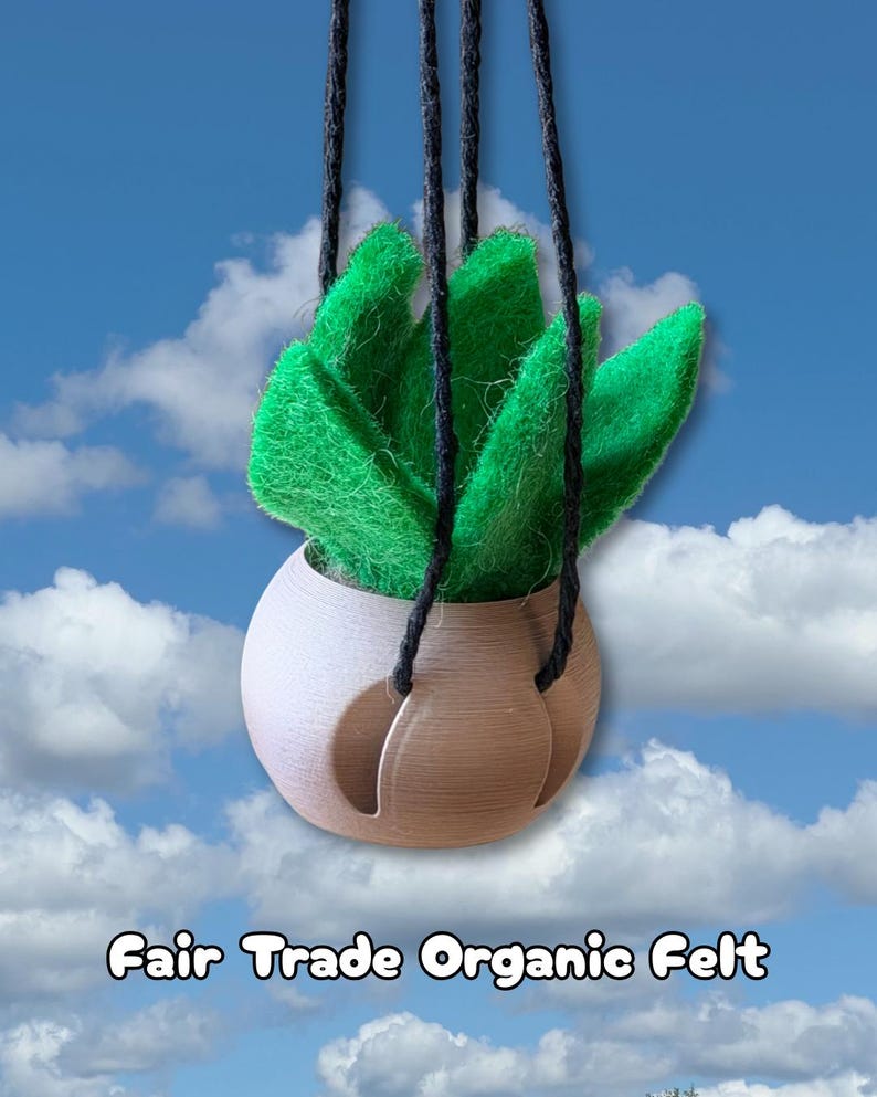 May include: A hanging plant decoration featuring bright green felt leaves arranged in a rounded, tan-colored planter. The planter is suspended by three black cords. The text "Fair Trade Organic Felt" is printed at the bottom of the image.
